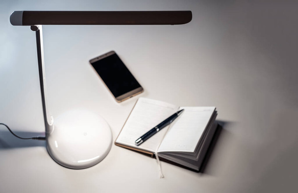 Egy okostelefon és nyílt napló egy tollal az asztalon az asztali LED lámpa. - Fotó, kép