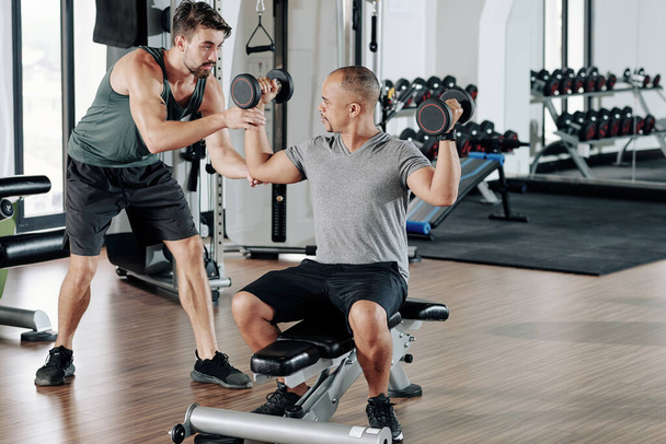Fitness coach explaining mixed-race young man how to work out with heavy dumbbells - Foto, Imagem