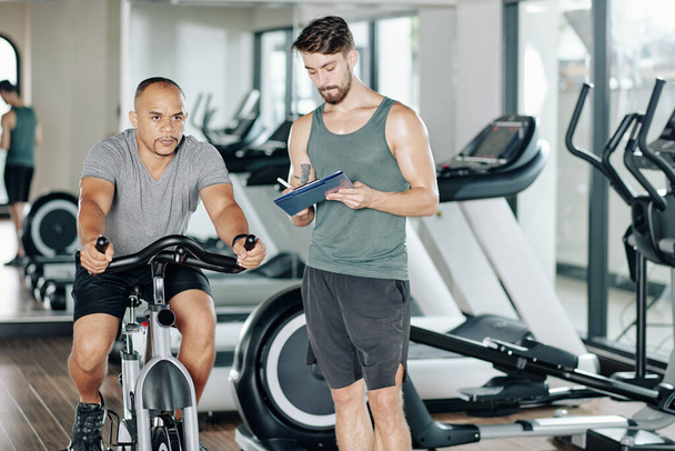 Fitness trainer writing in document when his client trying new cardio workout and riding fast on stationary bicycle - Foto, Imagen