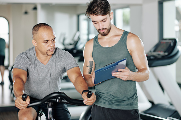 Hombre en forma discutiendo plan de entrenamiento con su entrenador al montar en bicicleta estacionaria en el gimnasio - Foto, imagen