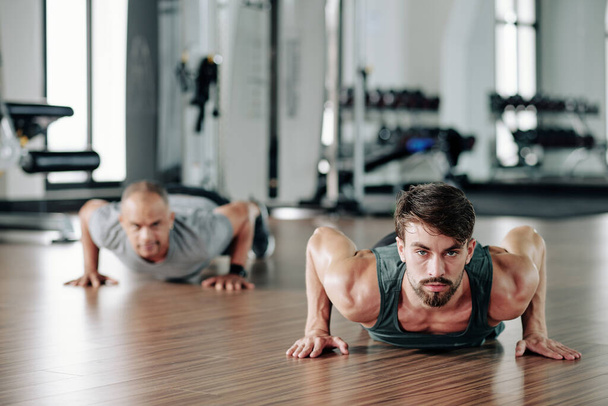 Handsome young fitness trainer showing half cobra push ups to mature client in gym - Photo, Image