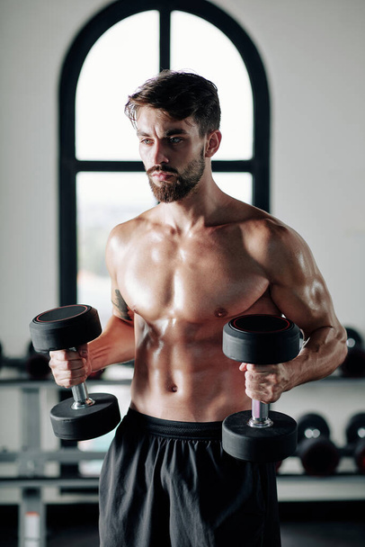 Erős, komoly, félmeztelen fiatalember gyakorol nehéz súlyzókkal az edzőteremben - Fotó, kép