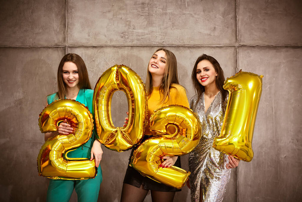 Glamorous young females holding shiny golden balloon numbers 2021 smiling at camera on gray wall - 写真・画像