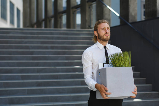 Nieszczęśliwy młody menadżer w białej koszuli zostaje zwolniony z pracy. Smutny pracownik siedzi na schodach centrum biznesowego po zwolnieniach. Kryzys i bezrobocie - Zdjęcie, obraz