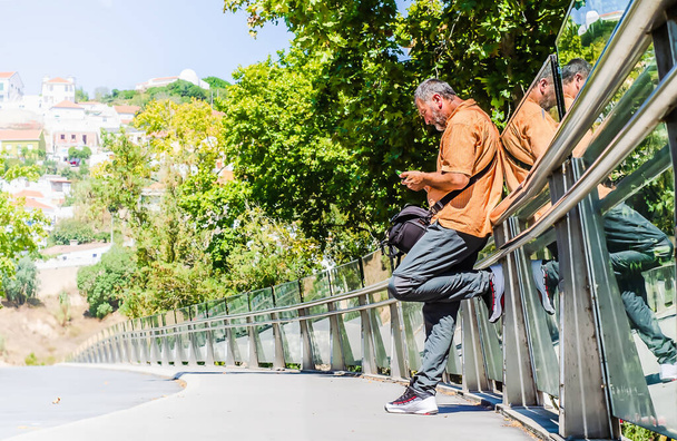 Ένας άντρας με πορτοκαλί πουκάμισο που ακουμπάει στο κιγκλίδωμα μιας γέφυρας συμβουλεύοντας τα μηνύματα του κινητού του. - Φωτογραφία, εικόνα