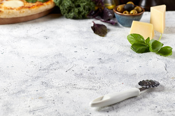 A closeup of a light gray wooden table with a pizza knife, on a background of spinach, cheese, olives and pizza. Template for food photography. - Фото, зображення
