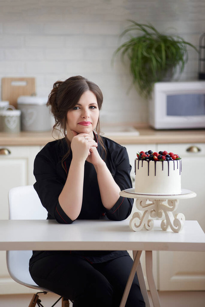 Confectioner young woman happy with her work sitting at table clasped hands looking at camera with beautiful juicy berry cake. Employment, freelancer at home concept, copy space. - Valokuva, kuva