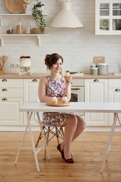 Young smiling woman sitting at the table, holding delicious cupcakes with cream and berries in white kitchen. Weight Loss Concept, copy space - 写真・画像