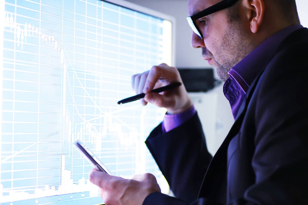 Ένας επιχειρηματίας κοιτάζει ένα γράφημα σε μια οθόνη. Ένας χρηματιστής αξιολογεί τις χρηματιστηριακές τάσεις. Ένας άνθρωπος με γυαλιά μπροστά από μια καμπύλη της δυναμικής της οικονομίας. - Φωτογραφία, εικόνα
