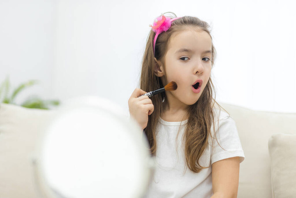 Photo of little cute girl holding cosmetic brush looking at the mirror on white background. - Fotoğraf, Görsel