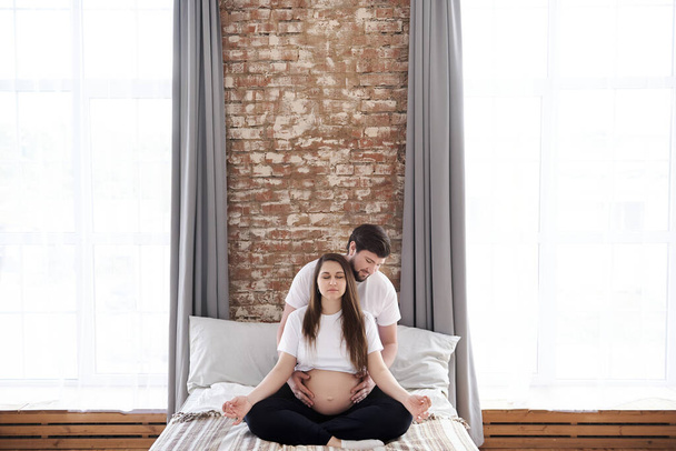 A pregnant woman sits in a lotus position with closed eyes and meditates on a bed while her husband gently hugs her. Being maternity and parenthood, wellbeing in family concept. Copy space, family - Photo, Image