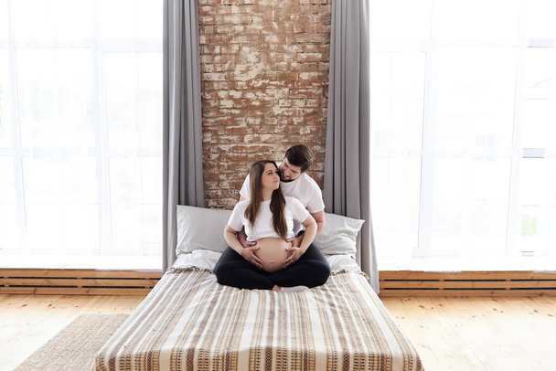Happy pregnant woman sits in a lotus position while her husband gently hugs and supports her on a bed at home. Pregnancy family time, healthcare, and wellbeing in family concept. Copy space. - Foto, Bild