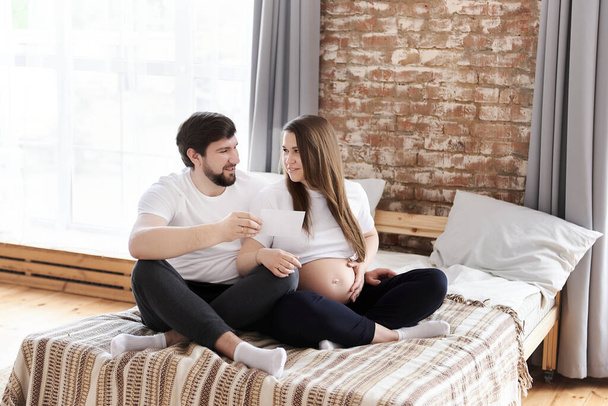 Happy young pregnant woman with her husband holding ultrasound image sitting on a bed at home. Being maternity and parenthood, wellbeing in family concept, pregnancy notice, special family moments - Photo, Image