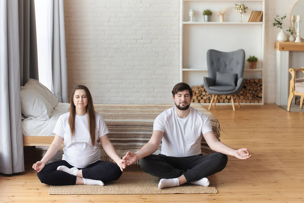 Young pregnant woman practicing yoga with her husband. They meditating in lotus position, their eyes closed. Pregnancy and being maternity and parenthood, wellbeing in family concept. Copy space. - Foto, Imagen