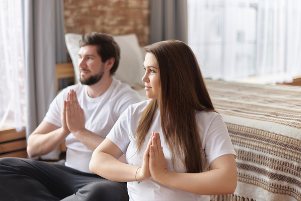 Young couple of beautiful pregnant woman and handsome husband practicing yoga and exercise, sitting with hands in Namaste and meditating, looking away at home, pilates training, copy space, close-up - Foto, Imagem