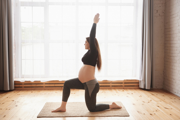 Young smiling pregnant woman, practicing yoga exercises at home or studio with big window. Prenatal variation of Warrior I posture. Pregnancy Yoga and Fitness concept. Full length, copy space. - 写真・画像