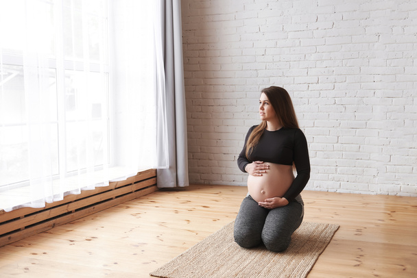 Young smiling pregnant woman sits on her knees a natural rug, doing hip stretching exercises, holds tummy, looks in the big window, rest after yoga exercise. Horizontal, Full length, copy space. - Фото, зображення