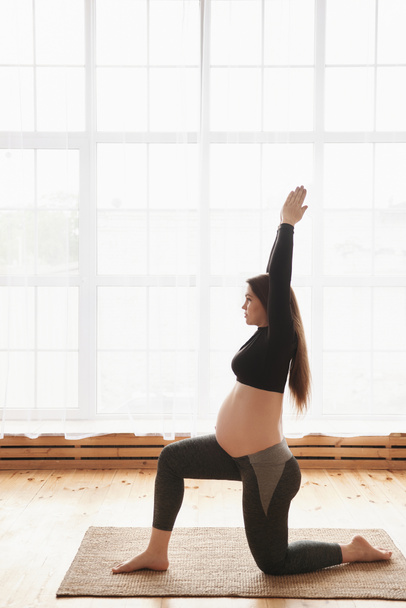 Young pregnant woman, practicing yoga exercises at home or studio with big window. Prenatal variation of Warrior I posture. Pregnancy Yoga and Fitness concept. Vertical, Full length, copy space - Foto, afbeelding