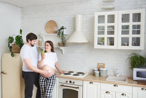 Future parents embrace standing in the kitchen. Pregnant wife gives husband a green apple. Healthy food, Lifestyle, parenthood, motherhood, expectation of a child. Copy space. Family pregnancy concept - Foto, imagen