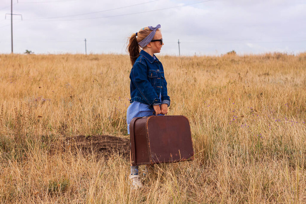 Little blonde girl in denim jacket, black sunglasses, blue dress with vintage suitcase in wild grass field. Stylish hitchhiker child with long hair countryside landscape trip. Kid walking outdoor road - Photo, Image