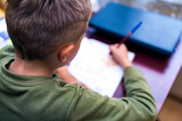 Läheltä katsottuna valkoihoinen koulupoika tekee läksyjä istuessaan työpöydän ääressä kotona. Kirjojen lukeminen ja oppituntien suorittaminen. Koulupoika tekee kotiläksynsä - koulutuksen käsite - Valokuva, kuva