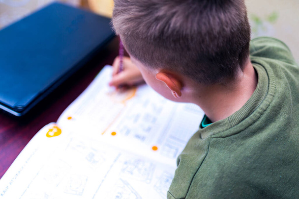 Läheltä katsottuna valkoihoinen koulupoika tekee läksyjä istuessaan työpöydän ääressä kotona. Kirjojen lukeminen ja oppituntien suorittaminen. Koulupoika tekee kotiläksynsä - koulutuksen käsite - Valokuva, kuva