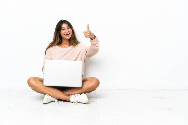 Young woman with a laptop sitting on the floor isolated on white background with thumbs up because something good has happened - Fotó, kép