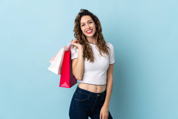 Young caucasian woman isolated on blue background holding shopping bags and smiling - 写真・画像