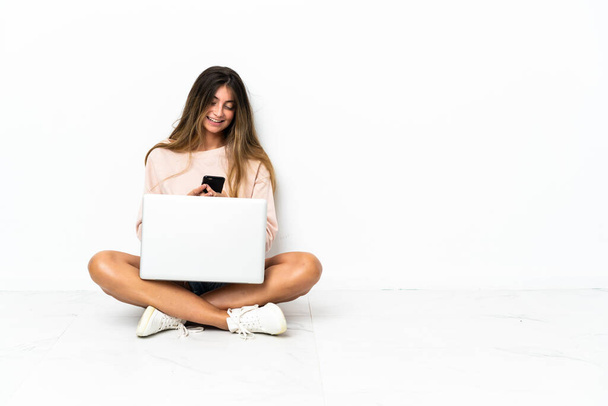 Молодая женщина с ноутбуком, сидящим на полу изолированы на белом фоне, отправляя сообщение с мобильного - Фото, изображение