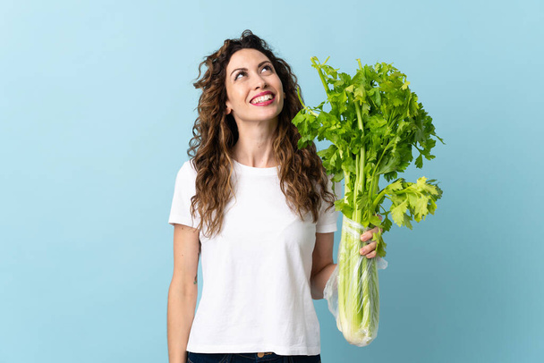 Young woman holding a celery isolated on blue background looking up while smiling - Photo, Image
