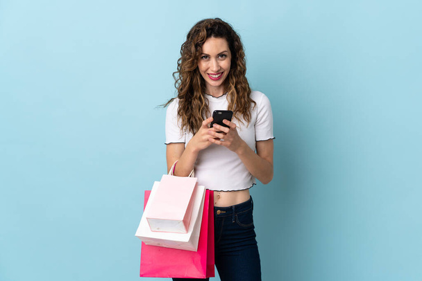 Giovane donna caucasica isolata su sfondo blu tenendo borse della spesa e scrivendo un messaggio con il suo cellulare ad un amico - Foto, immagini