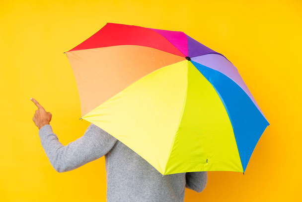 Hombre sosteniendo un paraguas sobre un fondo amarillo aislado apuntando hacia atrás con el dedo índice - Foto, imagen