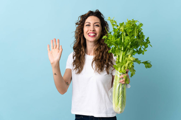 Young woman holding a celery isolated on blue background saluting with hand with happy expression - Photo, Image