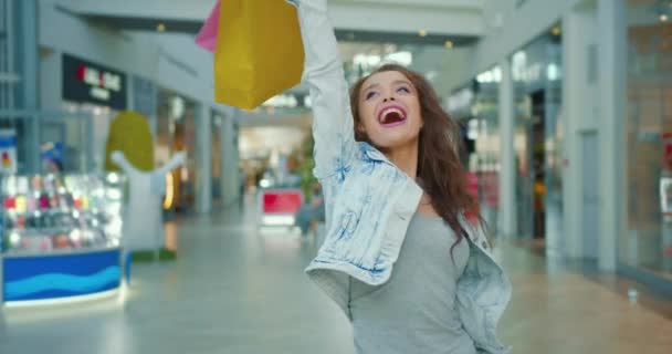 Het meisje is erg blij en glimlachend. Ze houdt de boodschappentassen omhoog. Winkels op de achtergrond. 4K - Video