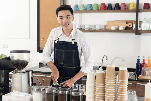 Улыбающийся азиатский бариста использует темпер для приготовления кофе для клиента, счастливо глядя в камеру в кофейне - Фото, изображение