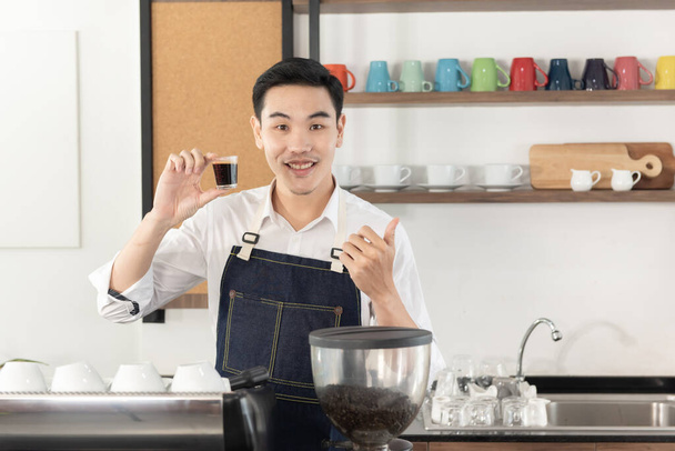 sonriente asiático barista hombre mostrar taza de negro café, golpes hasta sensación feliz mirada en cámara en café tienda - Foto, imagen
