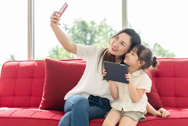 Šťastná asijská matka a malá dcera pořídit selfie foto smartphone, dívka drží tablet, úsměv a radostný, trávit čas s rodinou na holidě - Fotografie, Obrázek