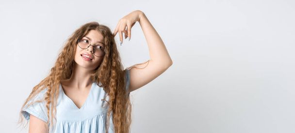 Photo of Pensive girl with curly long hair, and in round spectacles, looking up having pensive expression and holding her hand near head. Isolated over white background. Copy space for your text - Photo, image
