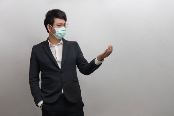 Asian businessman wear face mask and glasses with a white shirt and black suite show one hands, isolated on grey background - Фото, зображення