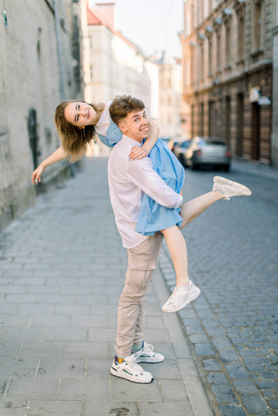Happy couple having fun outdoors in the city. Young handsome man holding his lovely girlfriend in blue dress in his arms outdoors, walking in the street - Foto, imagen