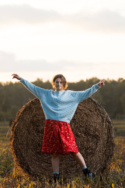 赤いドレスと編み物のセーターの美しい若いモデルの肖像画は、晴れた秋の日に干し草の上にポーズをとって、楽しいです。秋の暖かい写真。女性は笑顔と離れて見て、喜びの気分. - 写真・画像