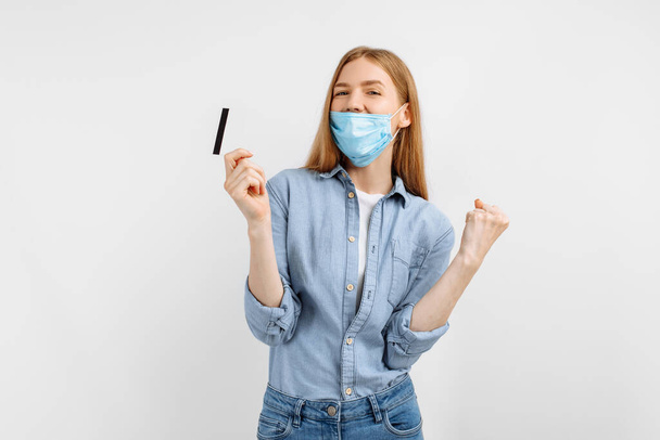 beautiful young woman in a medical protective mask on her face shows a winning gesture with a credit card in her hands on a white background - Foto, Imagem