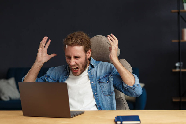 Angry young handsome curly man using laptop. Stressed freelancer lost data on broken not working laptop. Receiving bad news or email, spam message. Young people working with mobile devices - Foto, imagen
