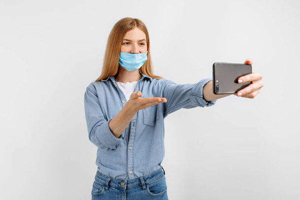 young woman in a medical protective mask on her face, talking on a mobile phone video link, on a white background, distance learning concept - 写真・画像