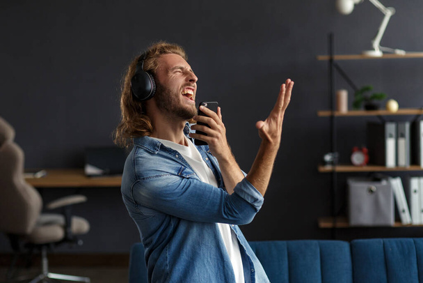 Handsome long haired curly man with headphones listening music,singing and dancing.Funny emotional smiling man with earphones and mobile phone relax at home.Enjoy listening to music.Stress management - Foto, Imagen