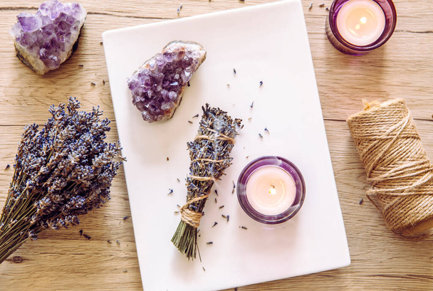 Homemade herbal lavender (lavendula) smudge stick smoldering on white plate with candles and amethyst crystal clusters for decoration. Spiritual home cleansing concept. - Foto, afbeelding