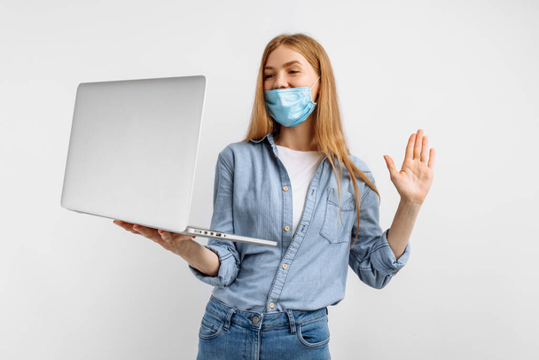 smart young woman in a medical protective mask on her face, talking on a laptop video link, on a white background, distance learning, working on the Internet - Фото, зображення