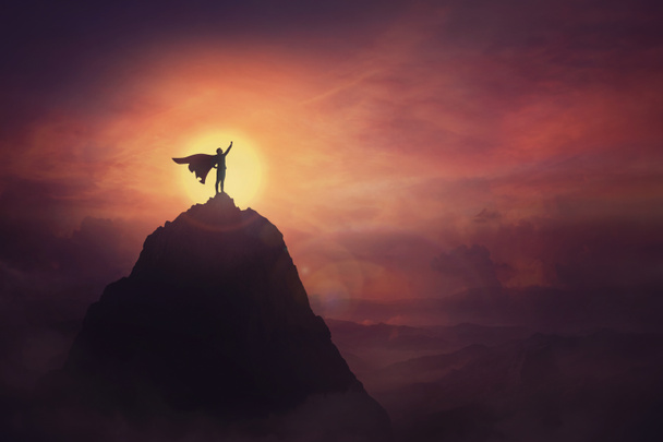 Conceptual sunset scene, superhero with cape standing brave on top of a mountain looks determined at horizon raising one hand up as a winning leader. Hero power and motivation, overcoming obstacles. - Photo, Image