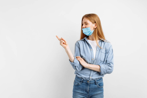 beautiful young woman in a medical mask on her face, points her finger at copyspace, on an isolated white background - Foto, imagen