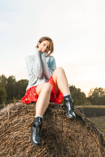 Porträt eines schönen jungen Models in rotem Kleid und Strickpullover, das an einem sonnigen Herbsttag auf Heuhaufen posiert. Herbstliches Foto. Frau lächelt und schaut weg, fröhliche Stimmung. - Foto, Bild